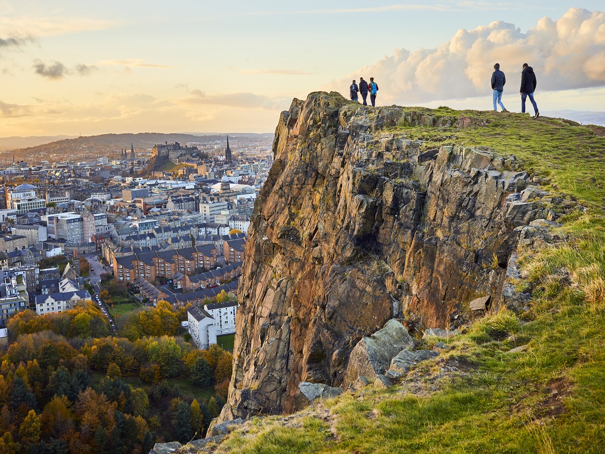 1058297160 Group of people walking along cliff edge looking at city views. Edinburgh Castle in the distance